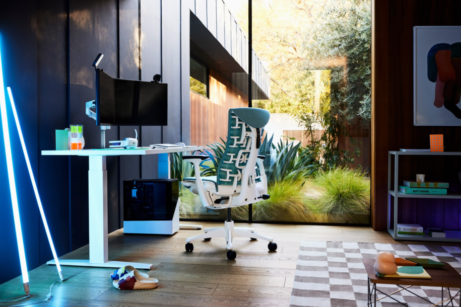 Desk inside of a room in front of window opening out to a courtyard with plants. The desk has two flat screen monitors and a white gaming PC on the floor beneath it.